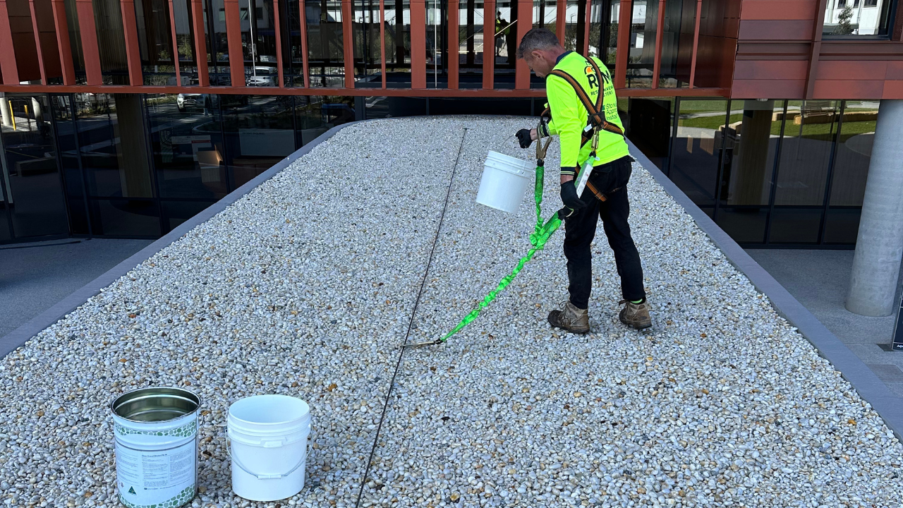 Installer in hi-vis and a safety harness spraying treatment across loose stone ballast on a hospital rooftop, with product containers in the foreground