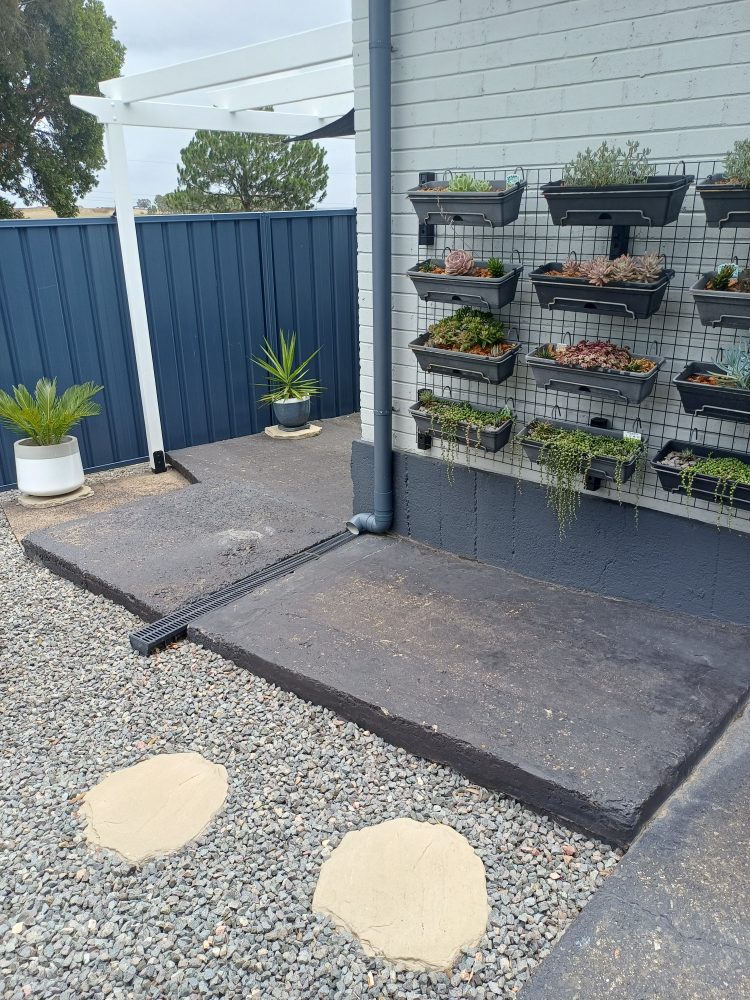Raised painted concrete slab beside a vertical garden on a dark fence, with a gravel area and round stepping stones in front, before resurfacing