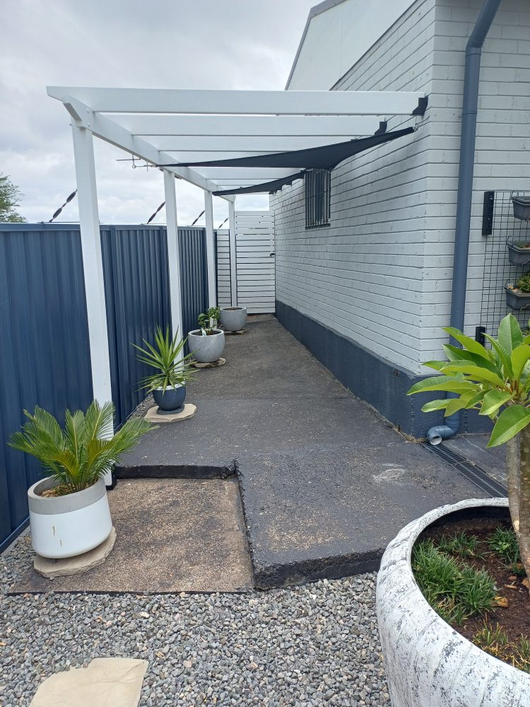 Outdoor area beside a house with worn, painted concrete under a white pergola, a dark fence, potted plants and gravel edging, before resurfacing