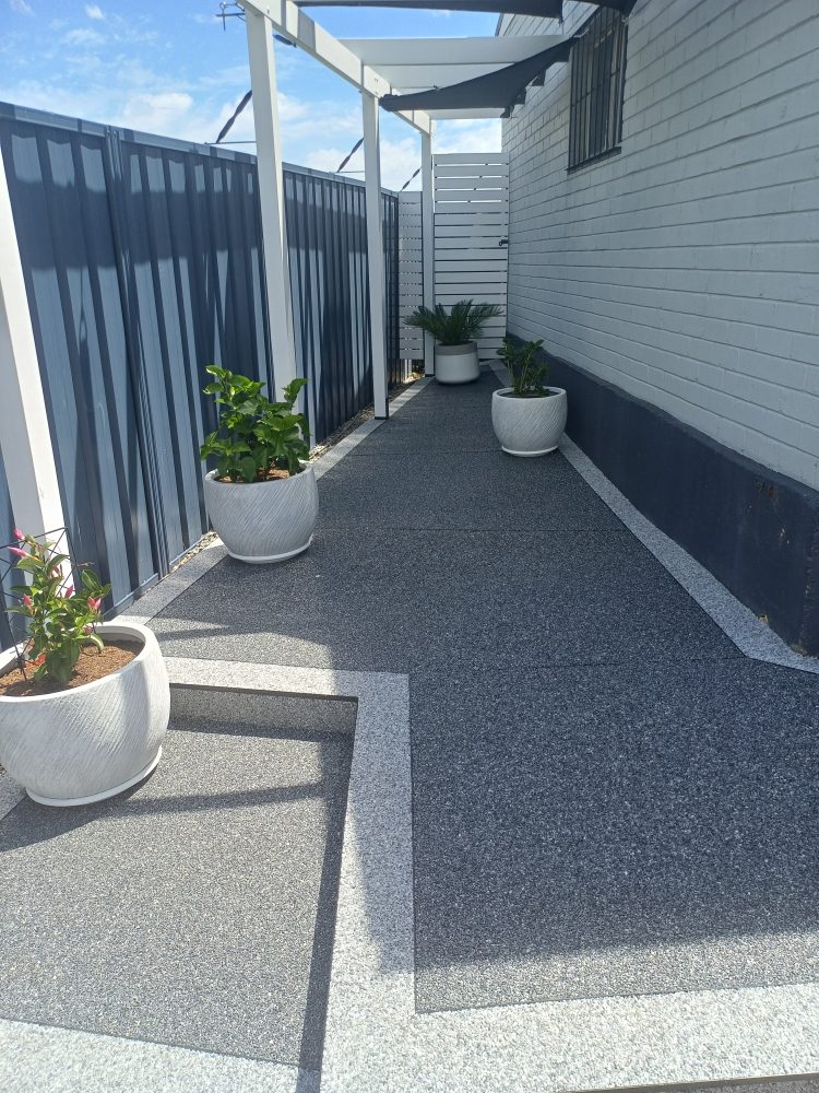 Outdoor area beside a house resurfaced in dark grey natural stone with a lighter grey border, under a white pergola, with potted plants and a dark fence